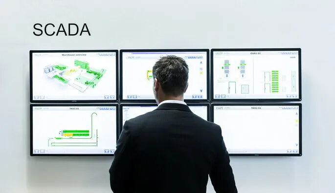 A person in a suit stands facing six large monitors displaying diagrams and data. The word SCADA is written on the wall to the left. The screens show various technical schematics and data charts.