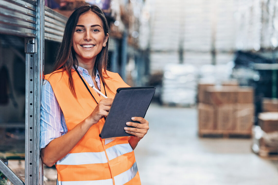 A woman in an orange safety vest stands in a warehouse, smiling as she holds a tablet. She leans against metal shelving, with stacked boxes and blurred storage racks hinting at the bustling world of online trading logistics in the background.