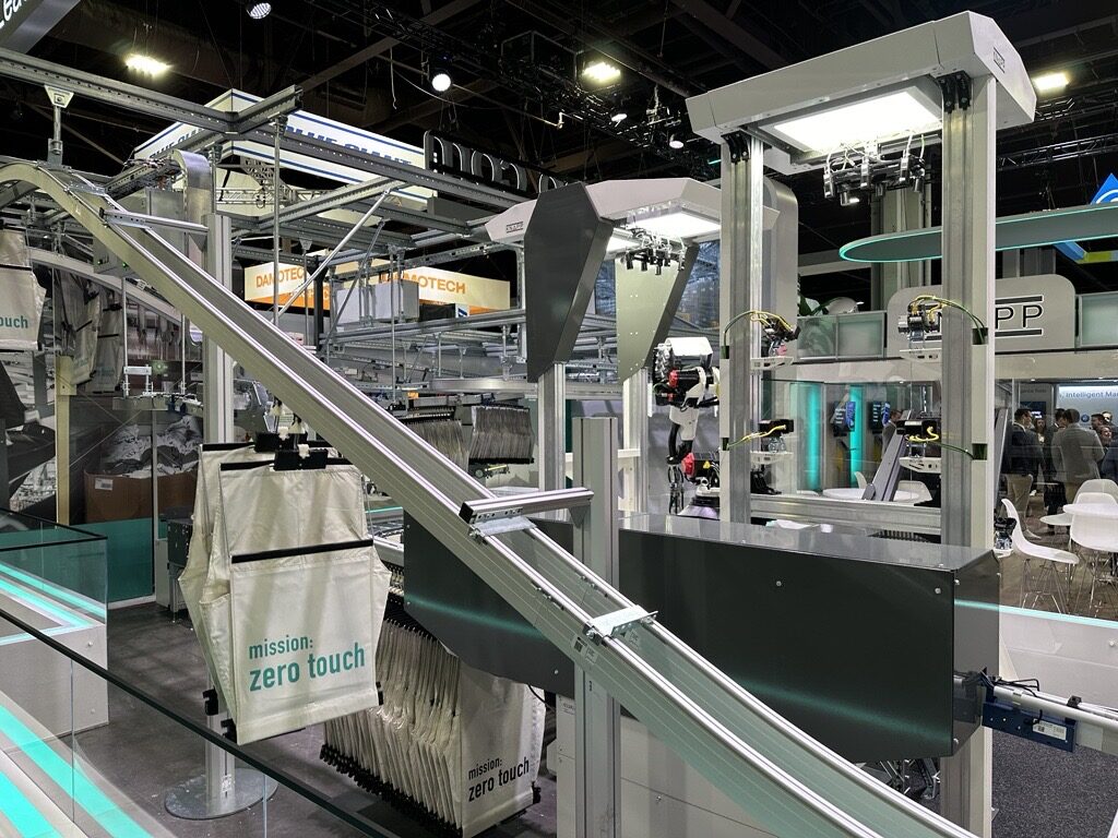 Advanced industrial machinery displayed at a tech exhibition, featuring a complex assembly line with robotic arms and conveyor belts. Banners read mission: zero touch, emphasizing automation. Booths and signage in the background.
