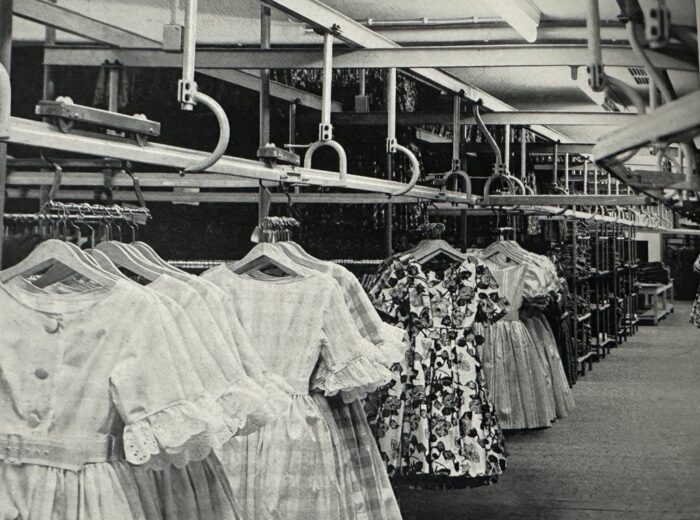 Black and white image of an automated garment conveyor system with dresses hanging on hangers. The dresses vary in patterns and are evenly spaced. The setting appears to be an industrial or factory environment.