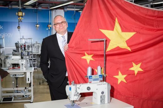 A man in a suit stands in a factory next to industrial sewing machines, smiling and holding up a large red flag with yellow stars. The background shows more sewing equipment.