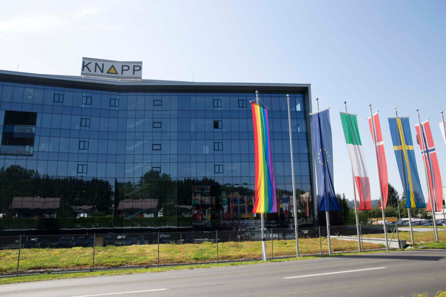 Modern glass building with KNAPP sign, surrounded by trees, and a line of nine flags including a rainbow pride flag, European Union flag, and various national flags like Italy, France, and Sweden, in front of a road.