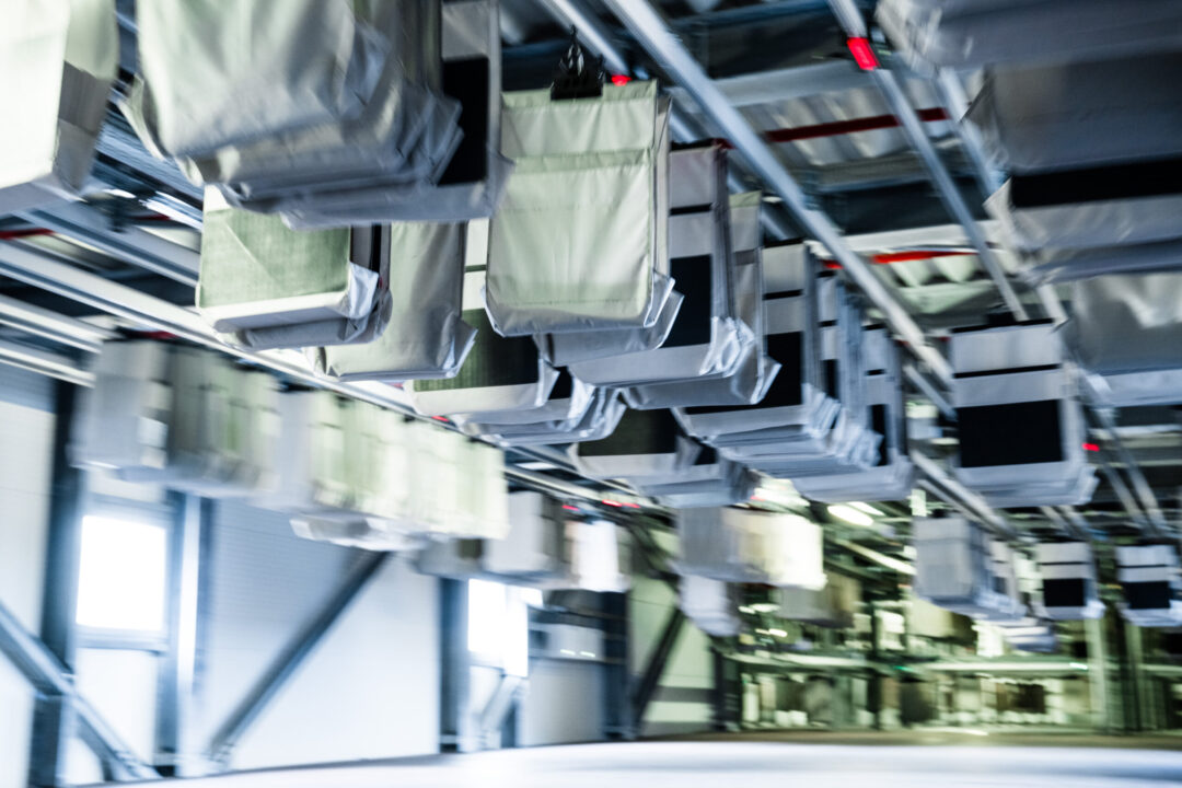 A blurred image of a logistical sorting system in a warehouse. Numerous fabric bags are attached to a network of rails suspended from the ceiling, used for transporting items. There are overhead lights and metal beams visible in the background.