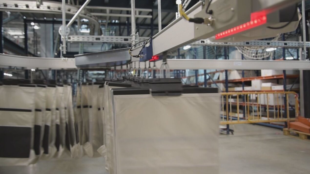 A conveyor system hanging from the ceiling transports large, beige fabric bags with black edges in a warehouse. The setup includes a network of rails and cables. The background shows shelves and a safety barrier.
