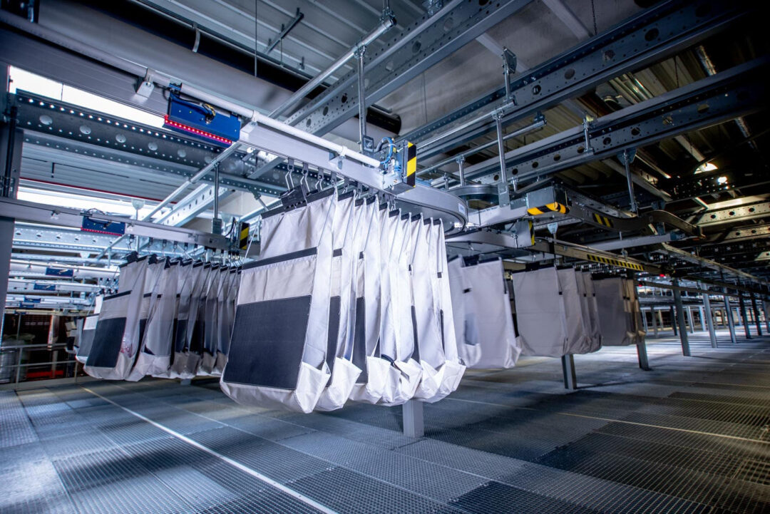 Rows of large grey fabric bags hanging from a metal rail system in an industrial setting. The scene features metal beams, a grated floor, and overhead conveyor belts, suggesting an automated storage or sorting facility.