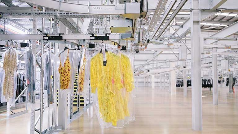 A modern garment warehouse interior showing clothes on hangers encased in plastic covers. Bright yellow patterned dresses are in the foreground, suspended on a conveyor system. The space is expansive, with metal beams and tracks overhead.