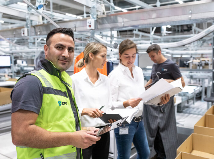 Ein Mann in einer Sicherheitsweste steht in einem Lagerhaus, hält ein Tablet in der Hand und lächelt in die Kamera. Er demonstriert effizientes Service Management, während drei Kollegen hinter ihm Dokumente prüfen und in der Nähe von Förderbändern und Kartons arbeiten.