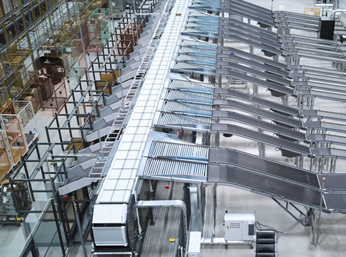 Ein großes Lager mit Push-Tray-Förderbändern und Sortierrutschen, die für die Handhabung und Verteilung von Paketen verwendet werden. In der geräumigen, gut organisierten Anlage sind Metallregale, Kisten und Industrieanlagen zu sehen.