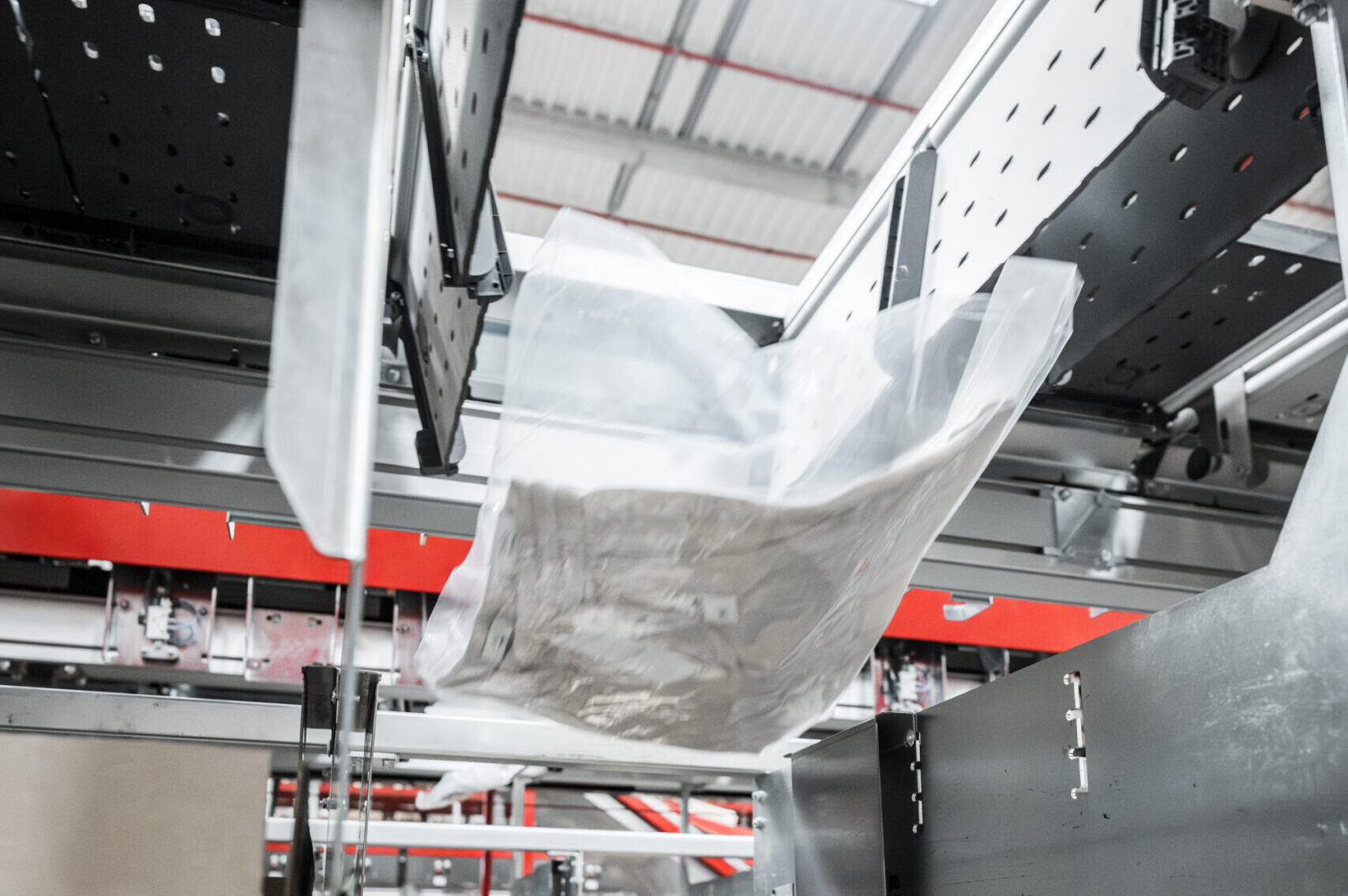 Ein durchsichtiger Plastikbeutel, der ein Paket enthält, bewegt sich entlang eines Split-Tray-Fördersystems in einer Industrieanlage mit Metallstrukturen und roten Akzenten.