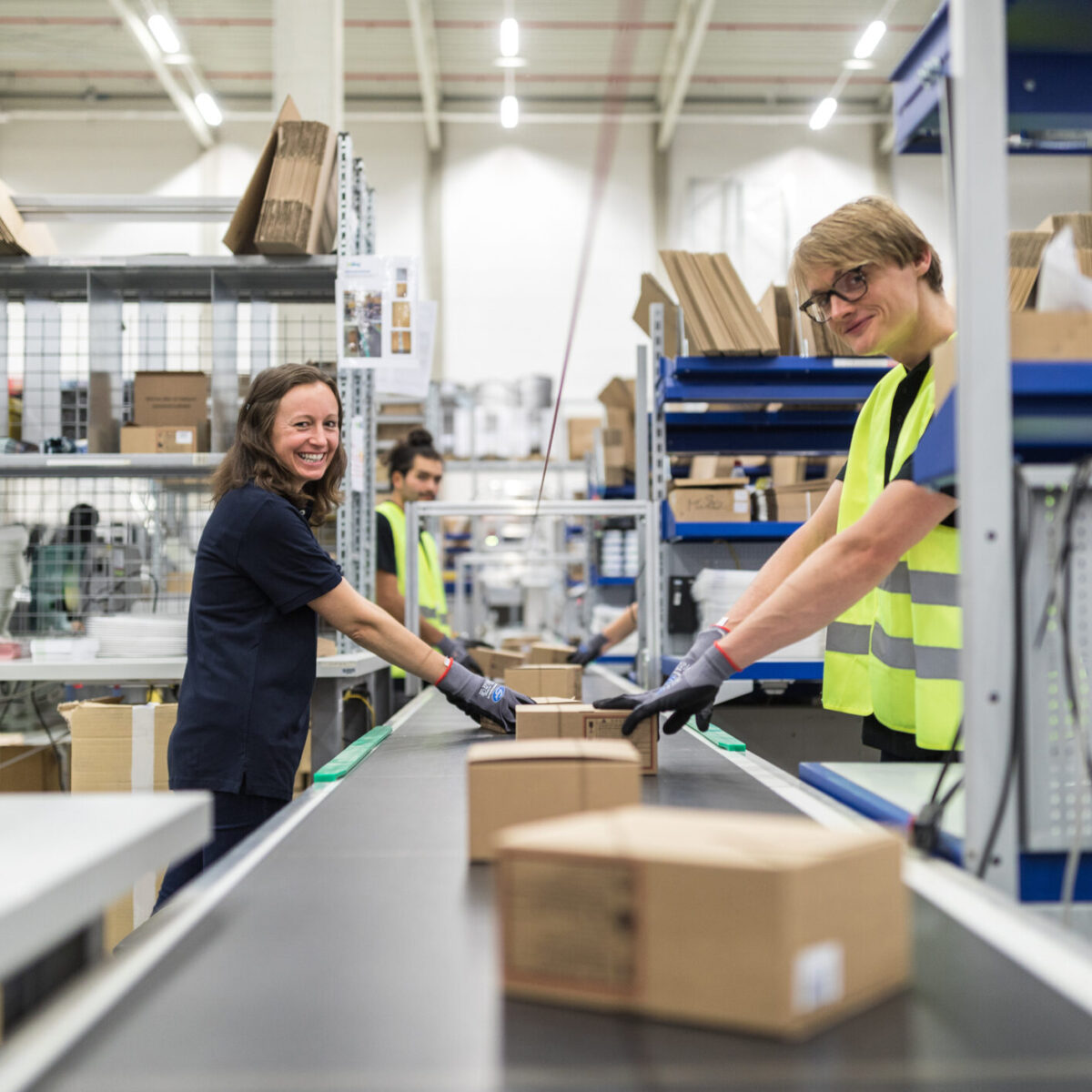 Two workers, one in a navy shirt and one in a yellow safety vest, smile as they handle returns and place cardboard boxes on a conveyor belt in a warehouse. Shelves with various items are visible in the background.