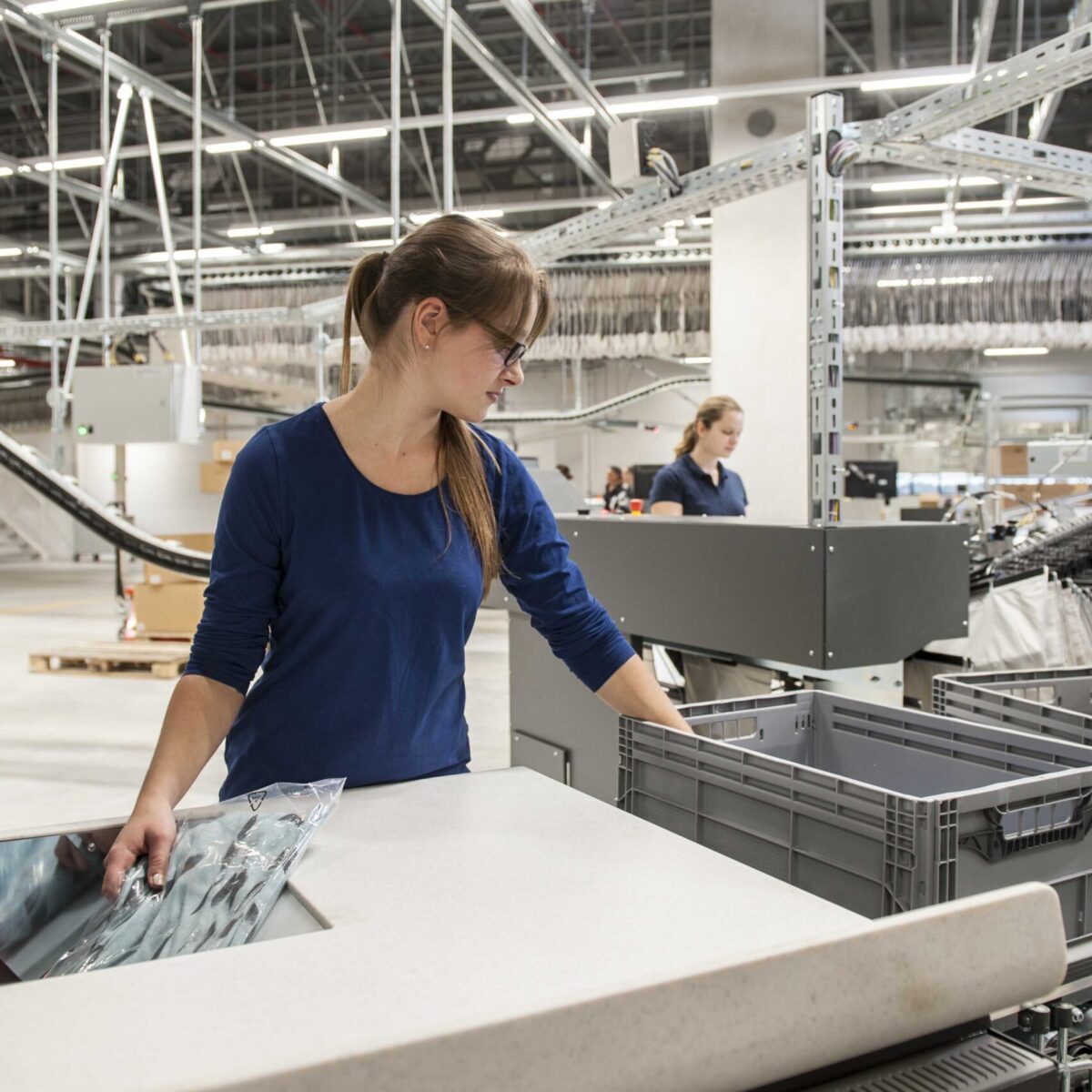 A woman wearing glasses and a blue shirt works at a modern industrial facility, focusing on returns management as she places packed items into a gray plastic bin on a conveyor belt. Another worker is visible in the background.