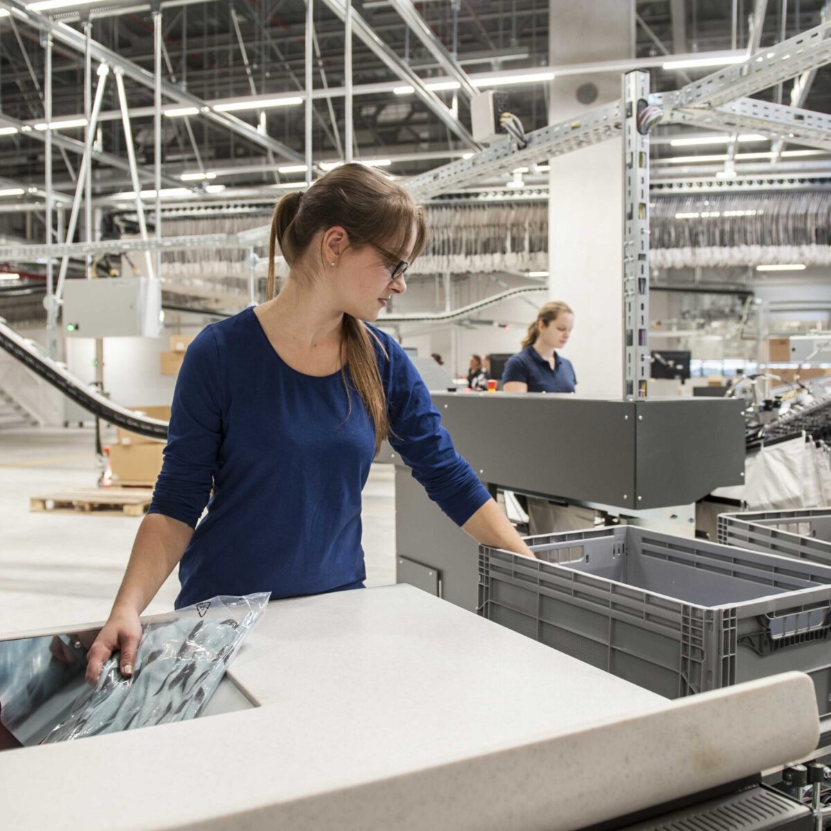 Eine Frau mit Brille und blauem Hemd hantiert mit verpackten Artikeln an einem Arbeitsplatz in einem modernen Lagerhaus, das sich auf Retourenmanagement konzentriert. Im Hintergrund sind ein weiterer Arbeiter und Industrieanlagen zu sehen.
