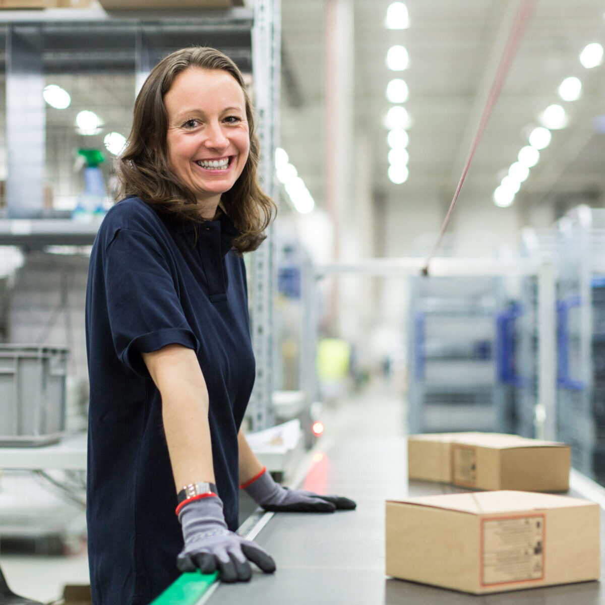 Eine lächelnde Frau in einem marineblauen Hemd und Handschuhen steht neben einem Förderband mit Kartons in einem Lager der Paketlogistik. Im Hintergrund sind Regale und Industrieanlagen zu sehen.