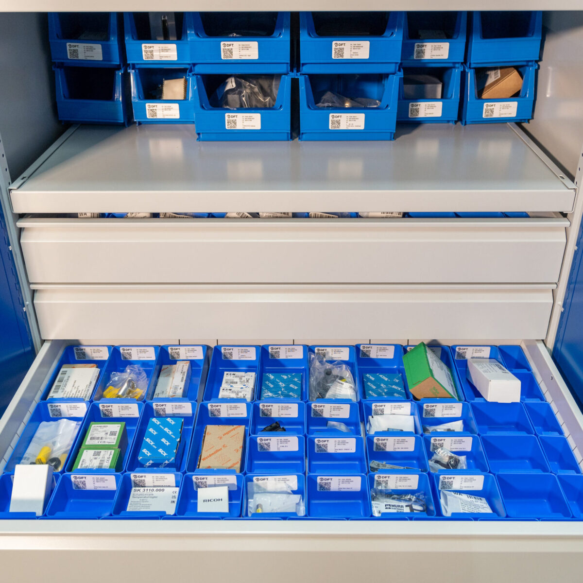 Blaue Plastikbehälter auf Regalen und in einer Schublade, die jeweils beschriftet sind und verschiedene Kleinteile oder Ersatzteilvorräte enthalten, wahrscheinlich in einem medizinischen oder industriellen Lagerschrank.