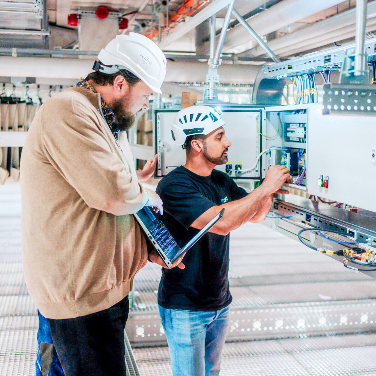 Zwei Ingenieure mit Schutzhelmen arbeiten gemeinsam an einer offenen elektrischen Schalttafel und unterstützen sich gegenseitig - einer hält einen Laptop, während der andere in einer hell erleuchteten Industrieumgebung die Verdrahtung im Inneren der Schalttafel justiert.