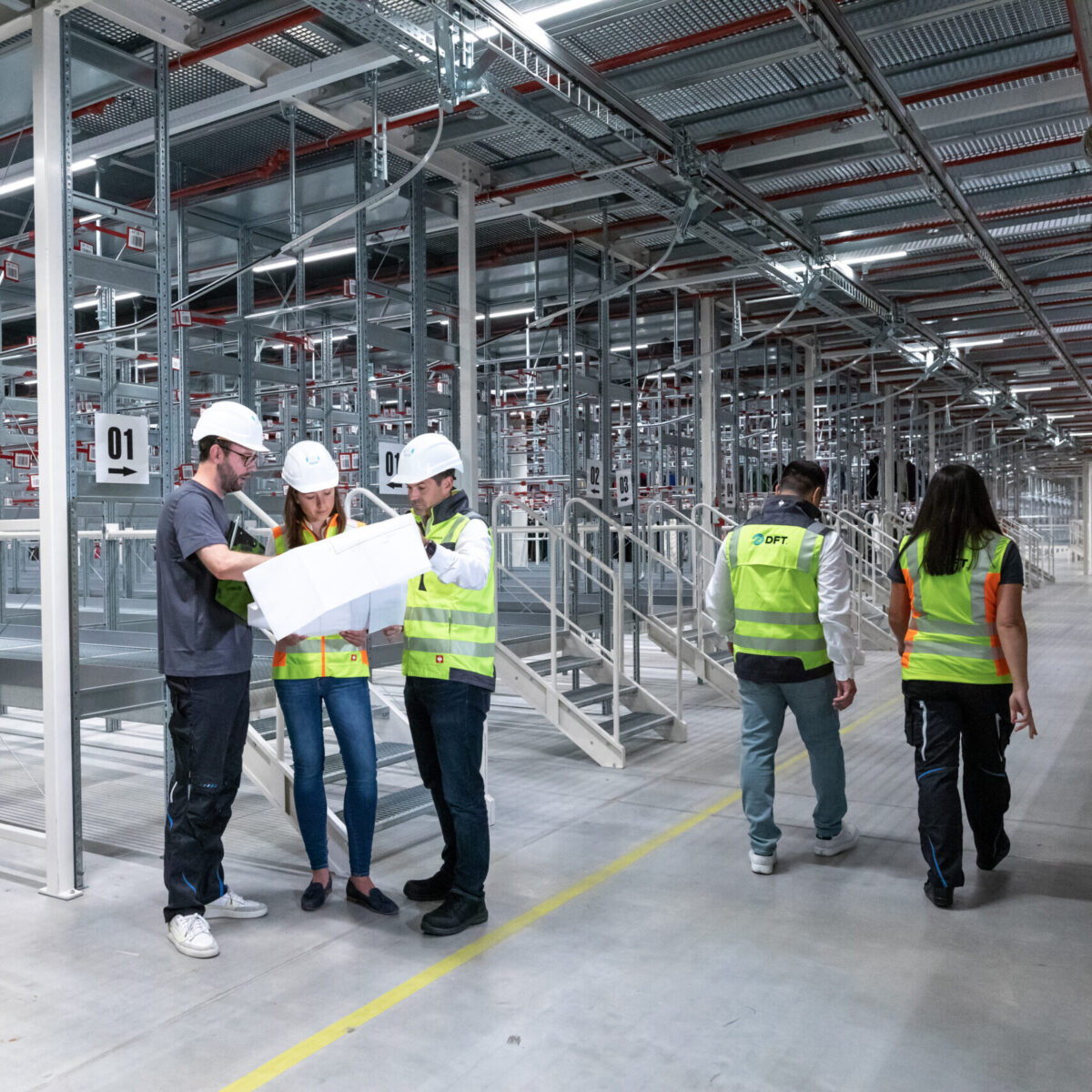 Four people in safety vests and hard hats examine blueprints in a large, empty industrial warehouse, possibly discussing recent training; two others walk away in the background between metal shelving units.