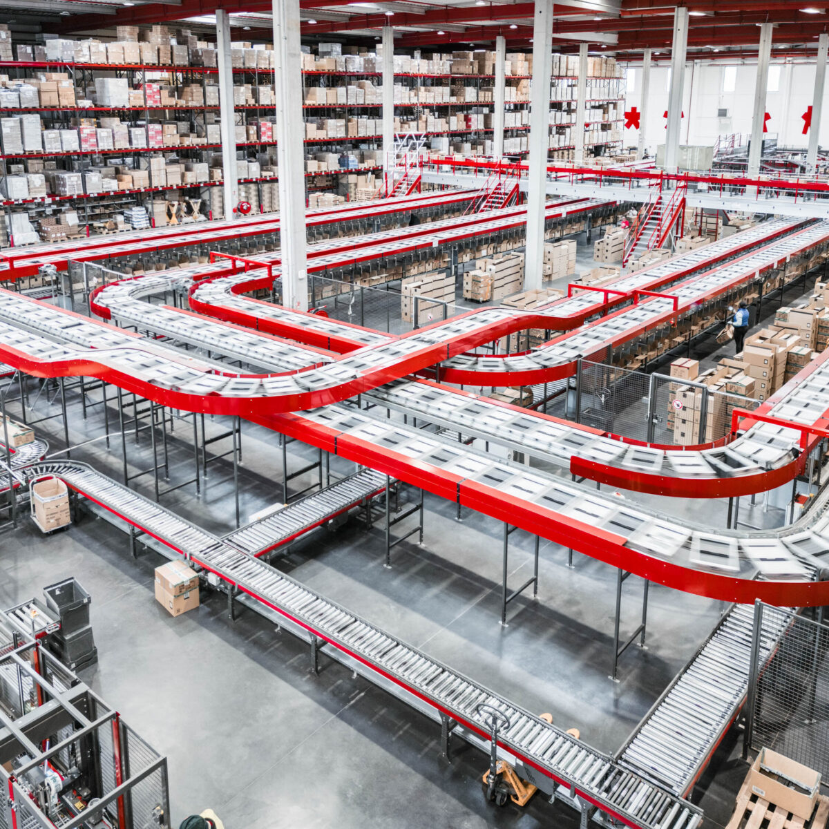 A large warehouse with tall shelves filled with boxes and a network of red and silver conveyor belts transporting store delivery packages across the spacious facility.