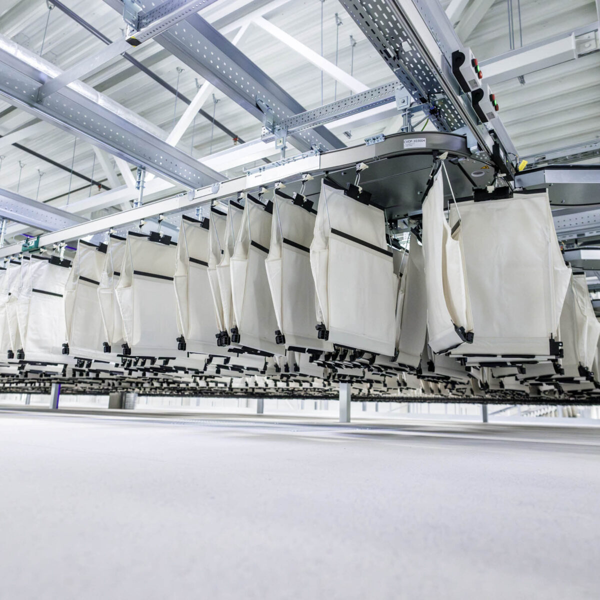 A row of large white fabric bags hangs from an automated conveyor system inside a modern pharmaceutical wholesale facility with exposed metal beams and bright lighting.