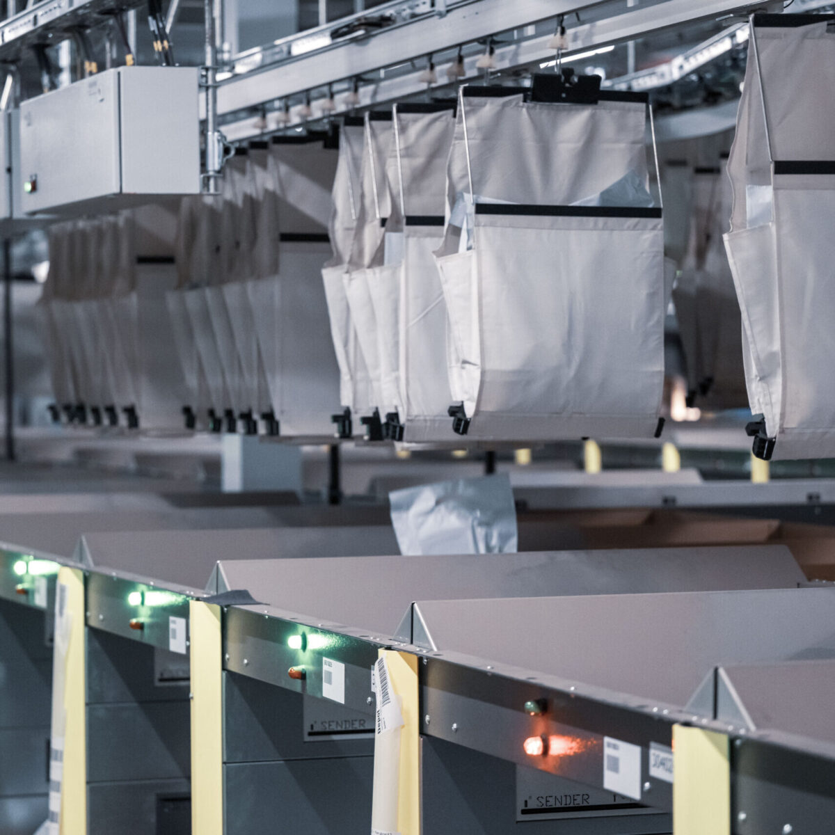 Cloth bags hang from an automated overhead rail system above conveyor belts with sorting bins and indicator lights, showcasing a modern industrial sorting facility as an efficient logistics solution.