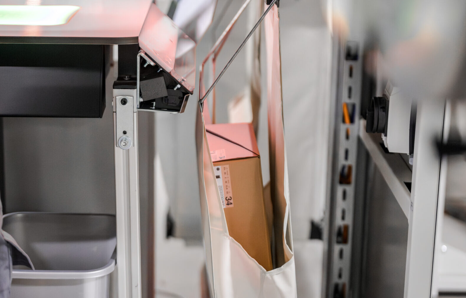 A cardboard box is suspended in an open cloth sling attached to a metal frame. The setup is positioned next to a desk with equipment and shelving visible in the background.