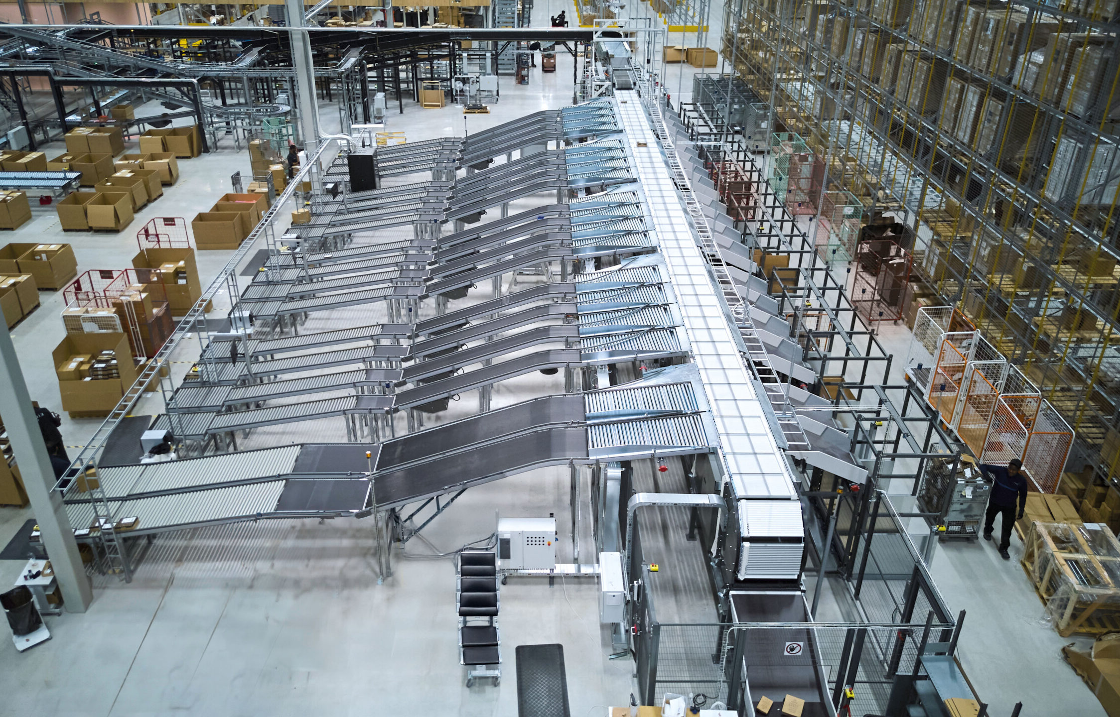 Ein großes Lager mit Förderbändern und Sortieranlagen für Pakete unterstützt die Ersatzteillogistik im Maschinenbau. Mit Kartons gefüllte Regale säumen die rechte Seite, während Arbeiter in der Nähe der Maschinen einen automatisierten Auftragsabwicklungsprozess hervorheben.