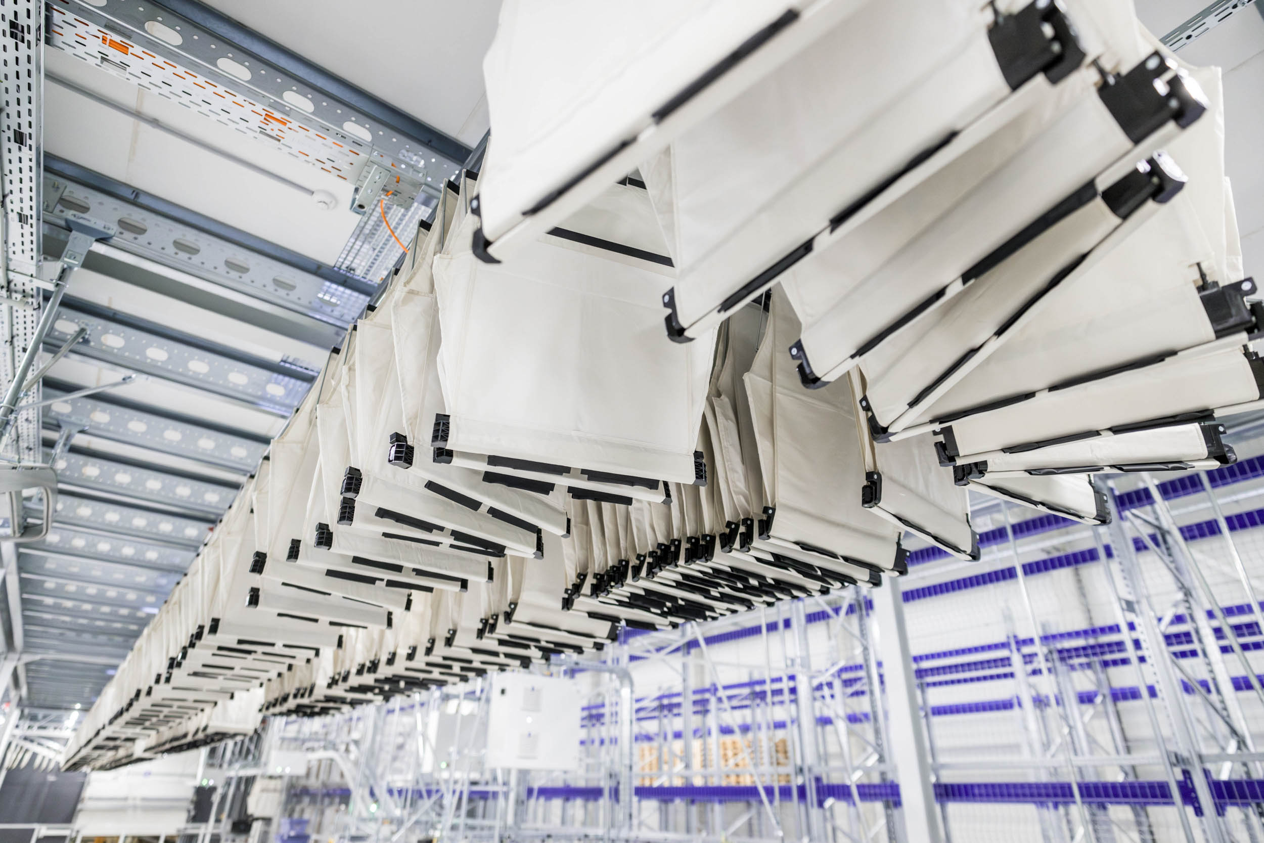 Reihen wei&szlig;er Stofftransporttaschen, die Teil eines Taschensortiersystems sind, h&auml;ngen an Metallschienen in einem automatisierten Lager oder einer Sortieranlage, im Hintergrund sind Regale und Lagergestelle zu sehen.